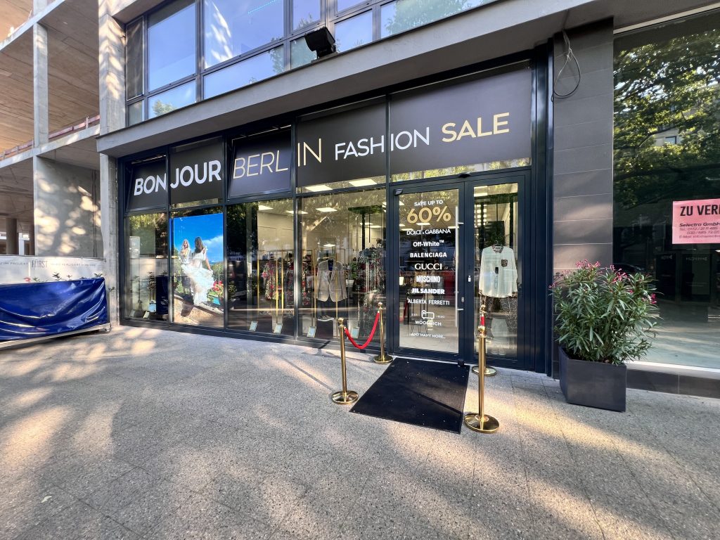 Außenansicht des Bonjour Berlin High Fashion Geschäfts. Das Schaufenster ist stilvoll dekoriert mit Designerstücken von Dolce & Gabbana. Die elegante Fassade zieht Kunden an und spiegelt den exklusiven Charakter des Geschäfts wider.